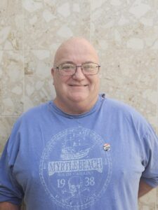 Older man poses and smiles in front of tan marble wall. He is wearing a blue t-short with "Myrtle Beach" text on it and Pride Pin.