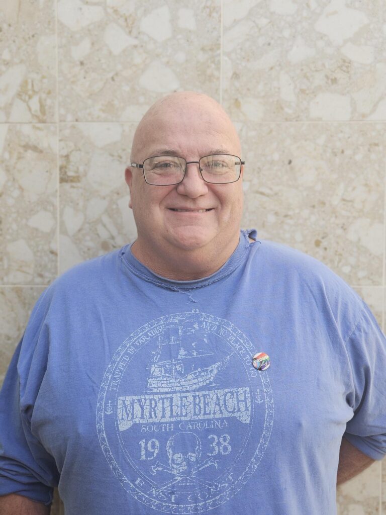 Older man poses and smiles in front of tan marble wall. He is wearing a blue t-short with "Myrtle Beach" text on it and Pride Pin.