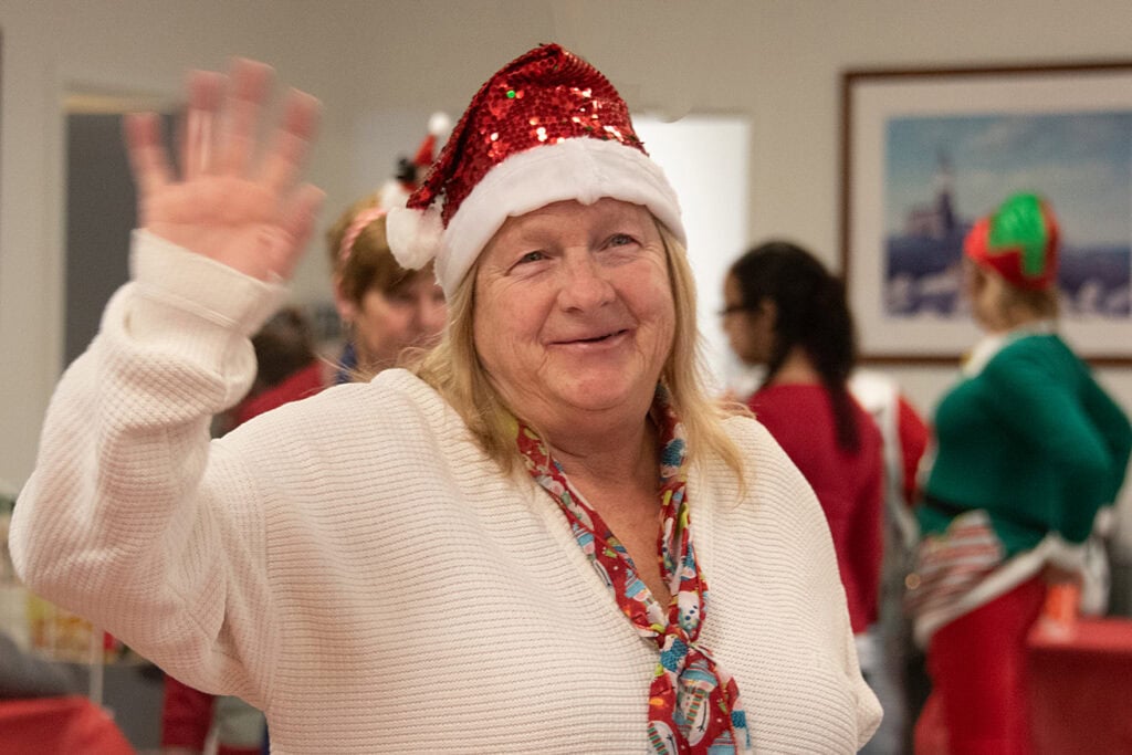 Shelter Coordinator, Bette, wearing her glittery Santa hat, waves a greeting at a holiday party.